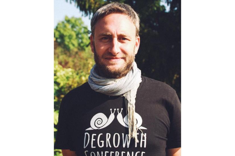 Photo of Vincent Liegey. Pictured smiling in a dark grey t-shirt which says &quot;Degrowth [...]&quot; and a light grey scarf. 